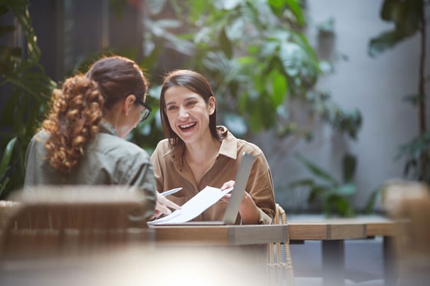 Two Smiling Young Women Discussing Travel in Cafe