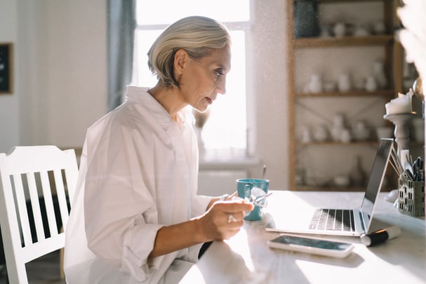 A woman at a kitchen counter with three devices in front of her—a phone, tablet, and laptop—looking thoughtfully at one of them, bright natural light, organized and calm energy