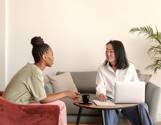 Two woman sitting one is working and the other one is looking at her to represent client and agent relationship