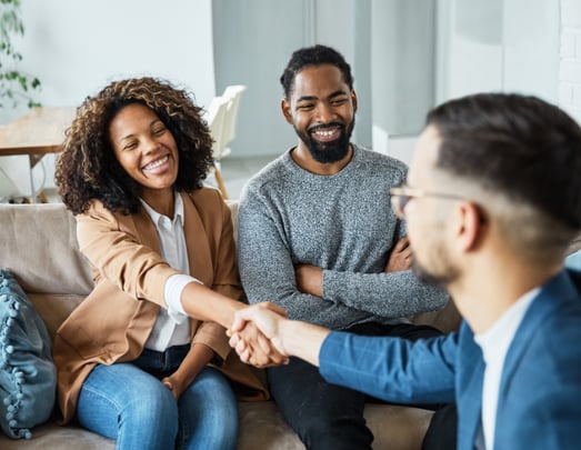In a bright, inviting home setting, a woman with curly hair, wearing a tan blazer and jeans, smiles broadly as she shakes hands with a man wearing glasses and a blue blazer. A bearded man in a gray sweater sits next to the woman, smiling. 