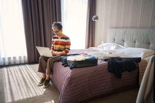 A stylish woman sitting cross-legged on a bed in a bright hotel room, editing photos on a laptop with a camera nearby, golden afternoon light, relaxed creative energy