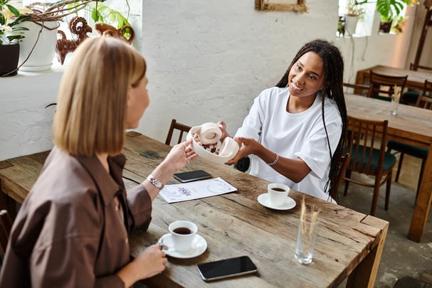 Two women sitting across from each other at a small café table, one leaning forward with attentive body language while the other gestures expressively, notebooks and coffee cups on the table