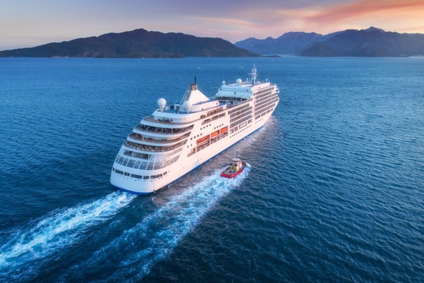 A large, white cruise ship sails on vibrant blue water in this high-angle shot, with a small red tugboat alongside. Numerous people are on the decks. Mountains are visible in the distance under a sky tinged with orange hues near the horizon.