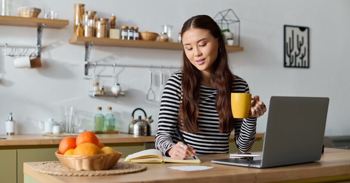 Woman working on a laptop at her kitchen table with a notebook and a cup of coffee