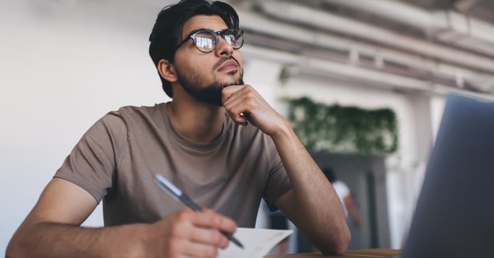 Thoughtful man writing notes with laptop nearby