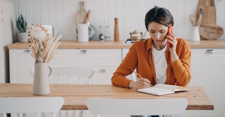 Focused woman taking notes and talking on mobile phone in bright kitchen