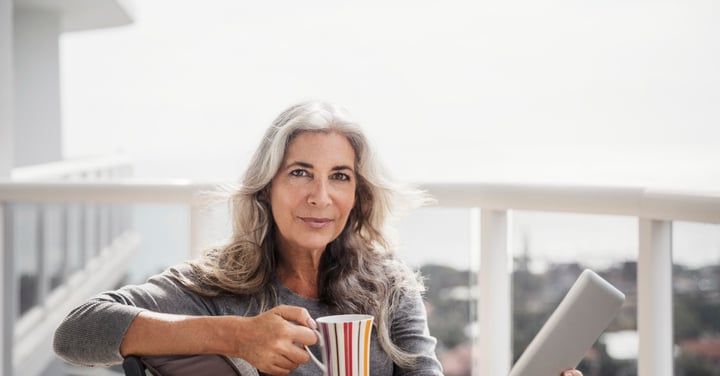 Senior woman holding coffee and tablet on sunlit balcony