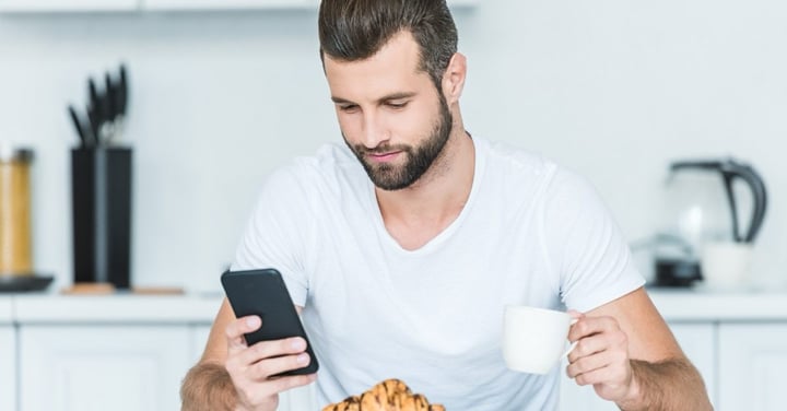 Smiling man reading a message on his phone over coffee and a croissant in a bright kitchen