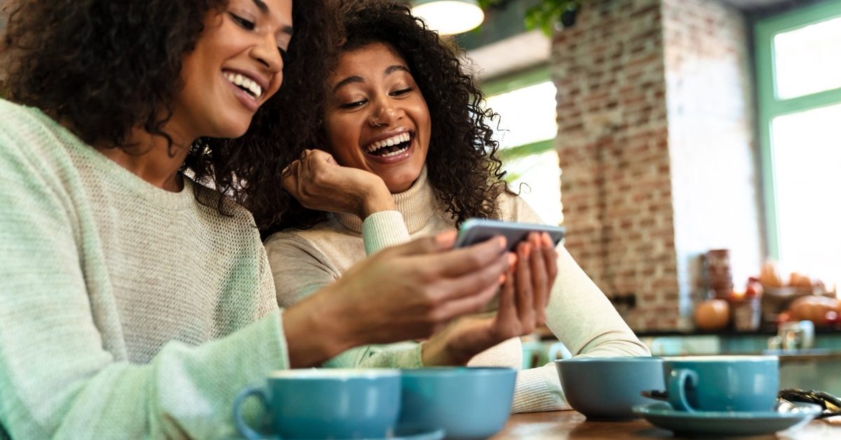 Two friends laughing together while looking at a smartphone over coffee at a cafe