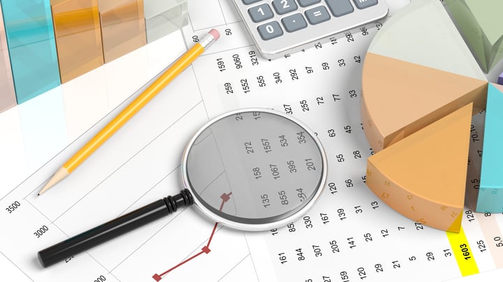 This overhead shot presents an arrangement of finance-related items on a white background. An open file shows a spreadsheet with figures and red lines. A three-dimensional pie chart in pastel colors lies to the side. A black magnifying glass is placed to the left, and a yellow pencil and grey calculator complete the composition. 