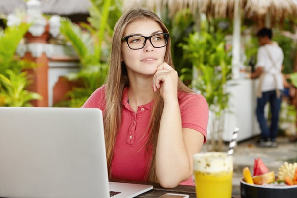 A woman with long, straight brown hair is shown seated at a table at an outdoor cafe. She wears glasses and a pink shirt and rests her chin on her hand, looking thoughtfully off to the side. A laptop is in front of her. A beverage and a bowl of sliced fruit are on the table next to her. The photo is sunlit and peaceful.